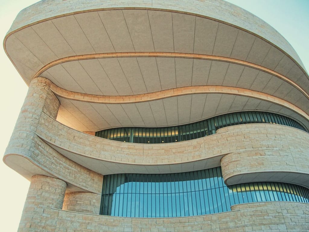 Curved architectural facade of a modern building with layered stone work and expansive glass windows under a clear, family-friendly sky.